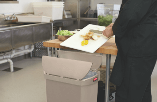 A Chef Scraping Food Waste Into A Pedal Bin Located Next To A Food Preparation Bench