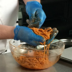 Chef Chris John wears blue gloves to lift orange spaghetti out of a bowl