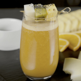 A glass of fruit juice beside a platter of chopped melon and oranges, topped with a skewer of melon, pineapple and kiwi